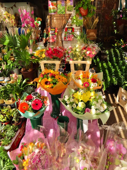 A vibrant display of fresh flower bouquets arranged on a pink cloth-covered table in a florist shop. The arrangements include bright red, yellow, pink, and white flowers such as daisies, roses, and ch
