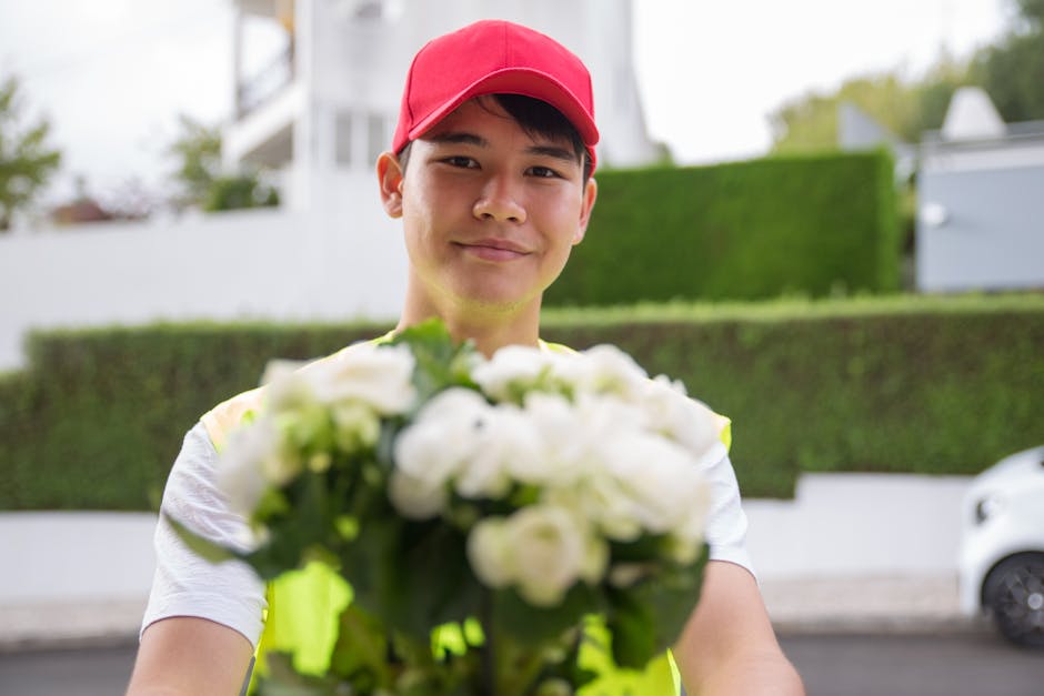 A young man wearing a red cap and a light-colored T-shirt is smiling as he hands over a bouquet of fresh white roses and greenery, wrapped in clear cellophane. The bouquet features tightly arranged pe