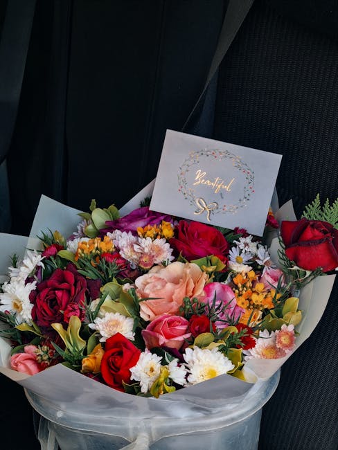 A vibrant bouquet of fresh flowers arranged in a clear wrapping, featuring red and pink roses, peach carnations, white daisies, and yellow filler flowers, with green foliage accents. The bouquet is re