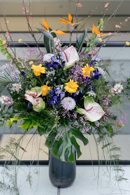 A tall floral arrangement featuring a variety of fresh flowers in a black ceramic vase, positioned on a windowsill with a blurred outdoor background. The bouquet includes bright yellow daisies, white