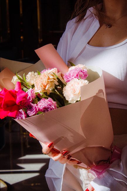 A person holding a bouquet of fresh flowers wrapped in light pink and white paper, featuring a mix of pink, white, and peach carnations, roses, and other blooms. The bouquet includes vibrant pink rose