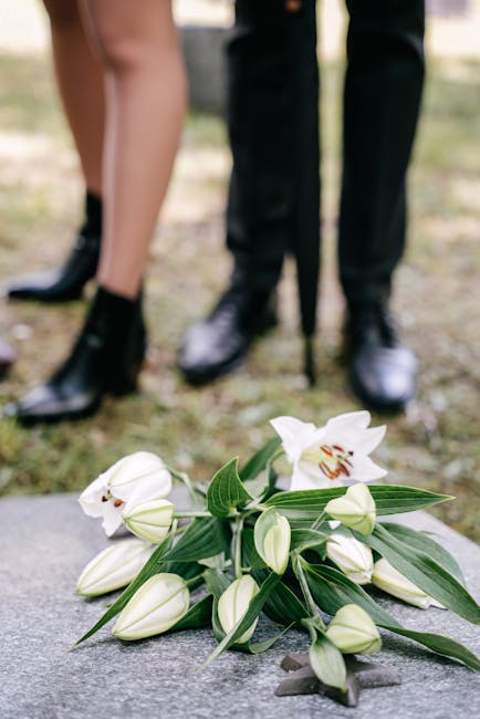 A bouquet of white lilies with open petals showcasing their reddish-brown stamens, surrounded by unopened lily buds and long green leaves, arranged on a stone surface. In the background, two people dr