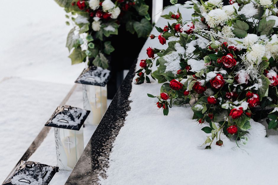 A floral arrangement featuring red roses and white flowers with green foliage, partially covered in snow, rests on a snow-covered surface. The flowers are arranged in a loose, natural style, with some